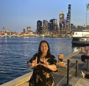 Dean Elizabeth Kronk Warner, a white woman with dark brown hair, positions her thumbs and forefingers to look like the letter "U" in honor of the University of Utah with the Chicago skyline in the background